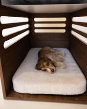 Dog lying on a fluffy cushion inside a wooden pet crate.