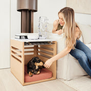 A dog is inside a wooden side table that doubles as crate furniture, with a person sitting on a bed reaching down to pet the dog. The room setting includes a lamp and a book on top of the side table.