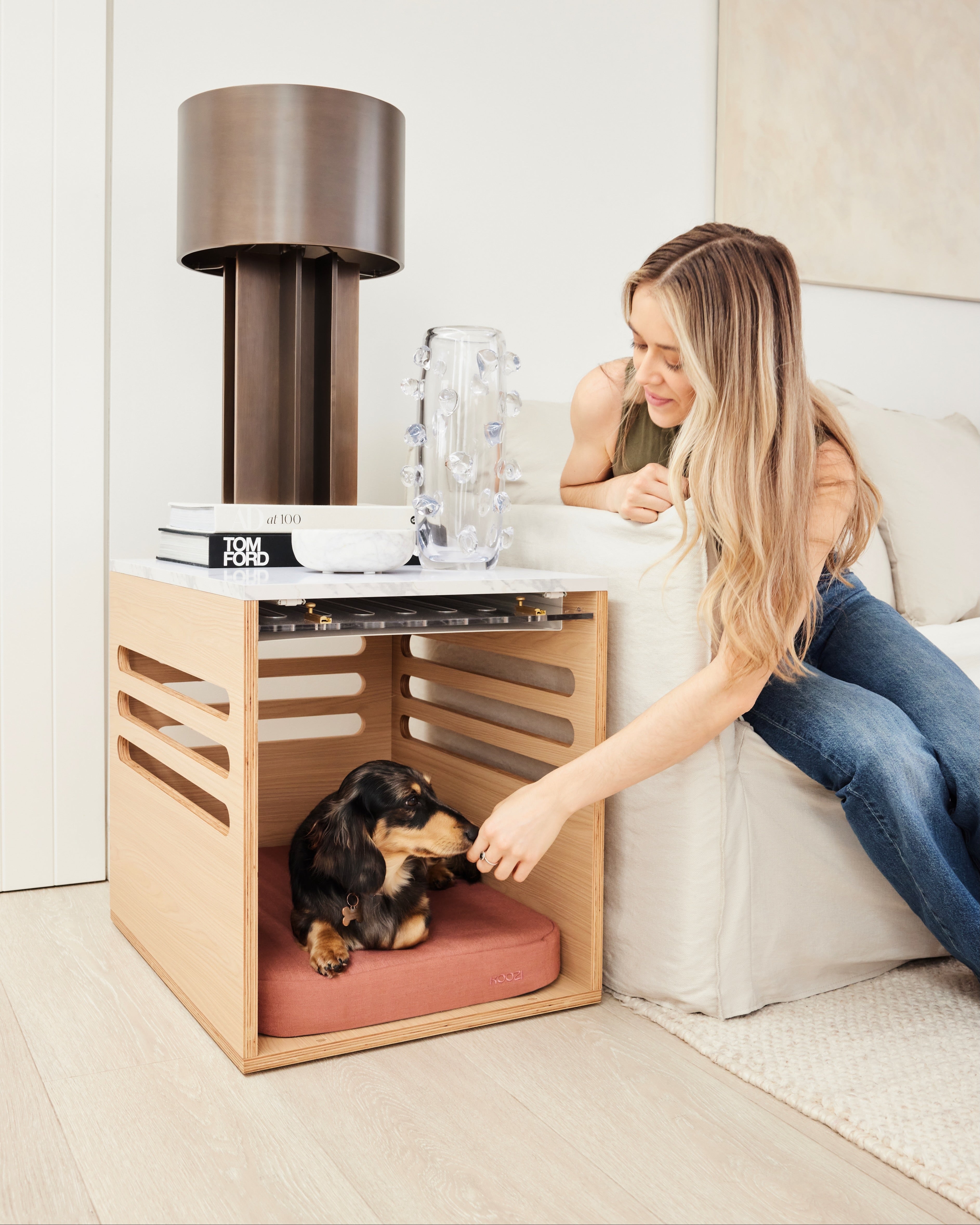 Woman petting a dog in a small wooden pet bed next to a lamp and books on a nightstand.